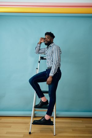 Black man in blue studio leaning on step ladder
