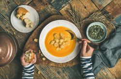 Top view of woman eating pumpkin soup with croutons, bread, seeds, pot, spoon on wooden table
