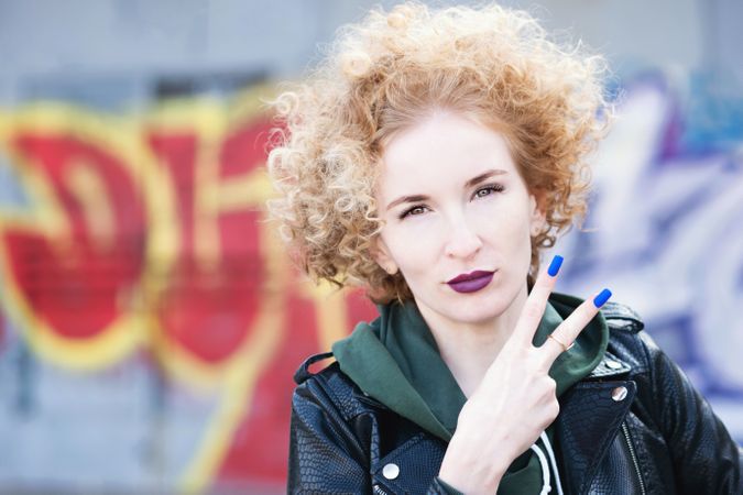 Curly haired female in leather jacket making peace sign outdoors