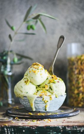 Bowl of melting pistachio ice cream with spoon, and grey background with leaves and glass jar