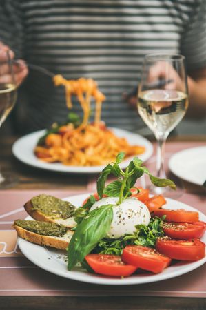 Man in striped shirt eating pasta with caprese salad in foreground, vertical composition