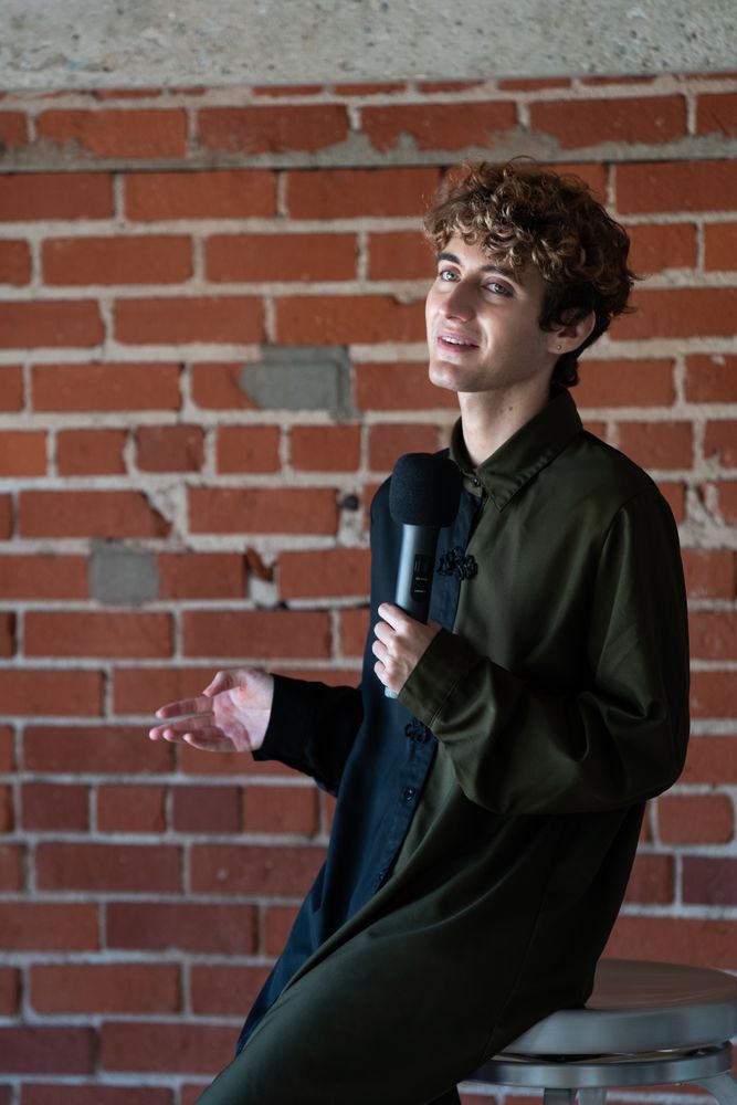 Nonbinary person giving talk holding microphone while perched on chair