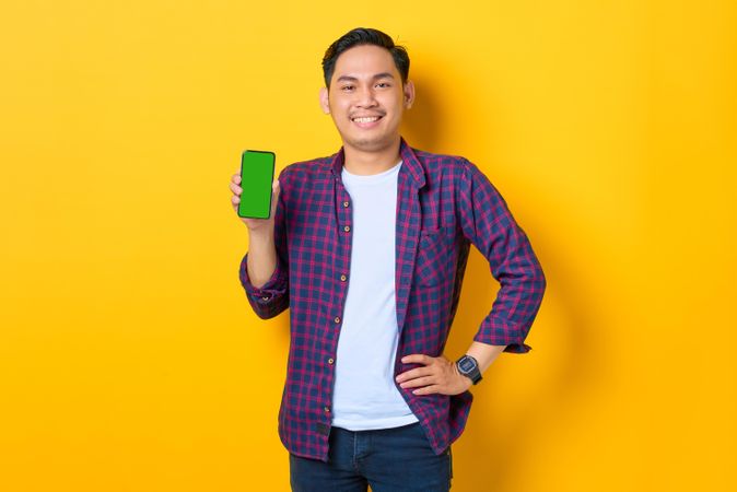Asian man with hand on hip in plaid shirt holding smartphone with chroma key