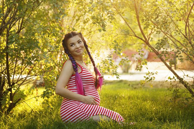 Pregnant female in red striped dress sitting under trees on the grass on sunny day