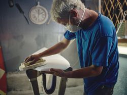 Male worker in respirator mask sanding surfboard with abrasive