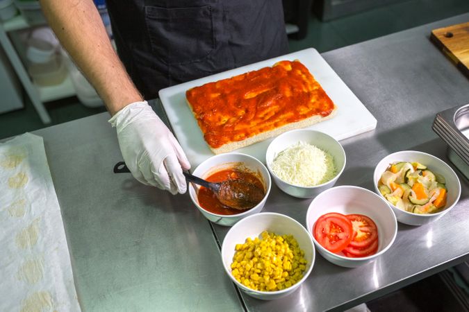 Unrecognizable male chef with gloves scooping tomato sauce to prepare pizza in restaurant kitchen