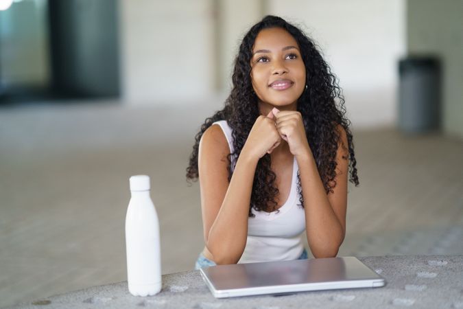 A thoughtful young woman with a laptop and a water bottle sits in a modern, stylish space