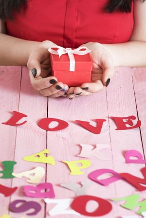 Woman’s hands holding a red gift box over a pink wooden table full of multicolor paper letters