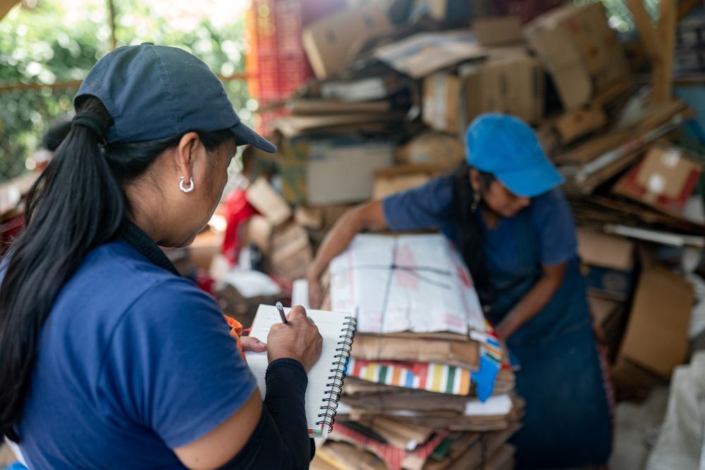 Hispanic Woman Noting Recycled Cardboard Inventory While Teammate Handles Stacks in Warehouse. Collaborative teamwork
