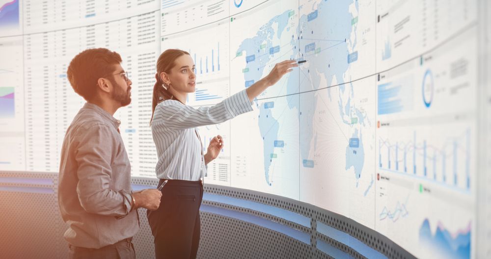 Indian Male Data Scientist And White Female Business Manager Talking In Front Of Big Digital Screen With Graphs, Charts And Map In Monitoring Office.