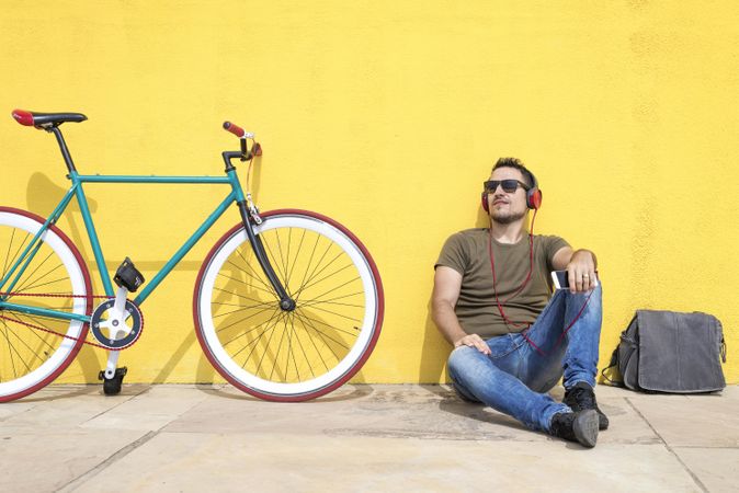 Male looking up in front of yellow wall next to bike with bike and listening to music