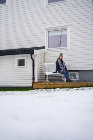 Thoughtful female tourist in warm clothing delighting in weather while resting on bench near house