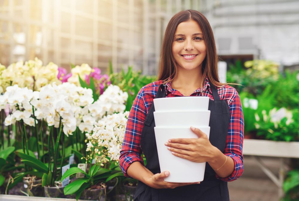 Smiling woman holding planters surrounded by flowers in a greenhouse