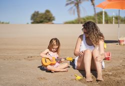 a little girl playing the ukulele sitting on the sand on the beach next to a mother