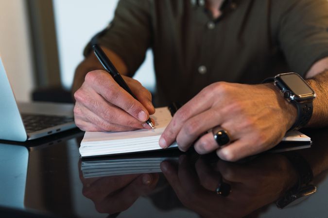 Anonymous man writing in notebook near laptop