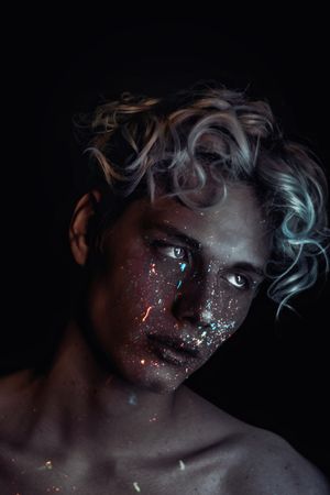 Portrait of topless blonde man with UV paint on his face looking away against dark background