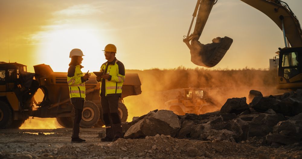 Cinematic Golden Hour Shot Of Construction Site: White Male Civil Engineer And Hispanic Female Architect Talking And Using Tablet. Trucks, Excavators, Loaders Working To Build New Apartment Block.