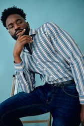 Black male model in striped blue shirt and jeans in blue studio leaning on step ladder