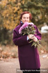 Older woman in purple coat holding bouquet of purple flowers standing near yellow trees 0vK6d5