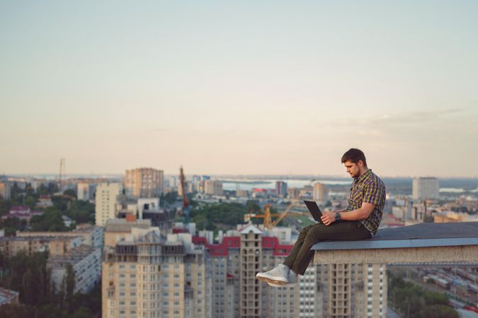 Side view of man perched on roof with legs dangling over city while working on laptop