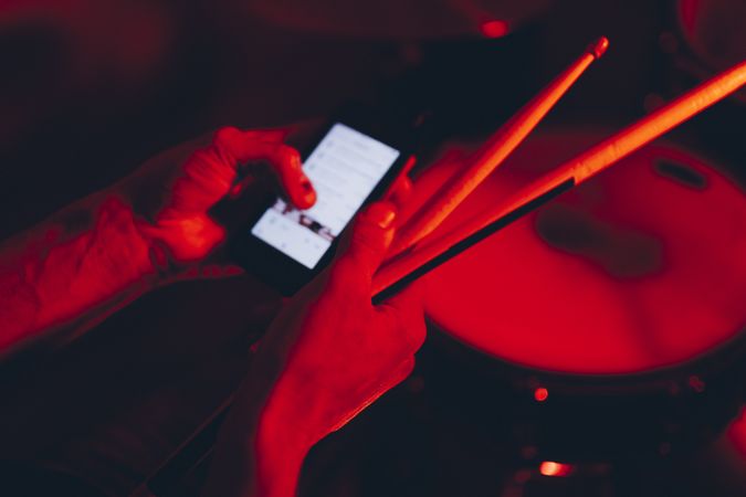 Man with drumsticks using smartphone near drums in redness