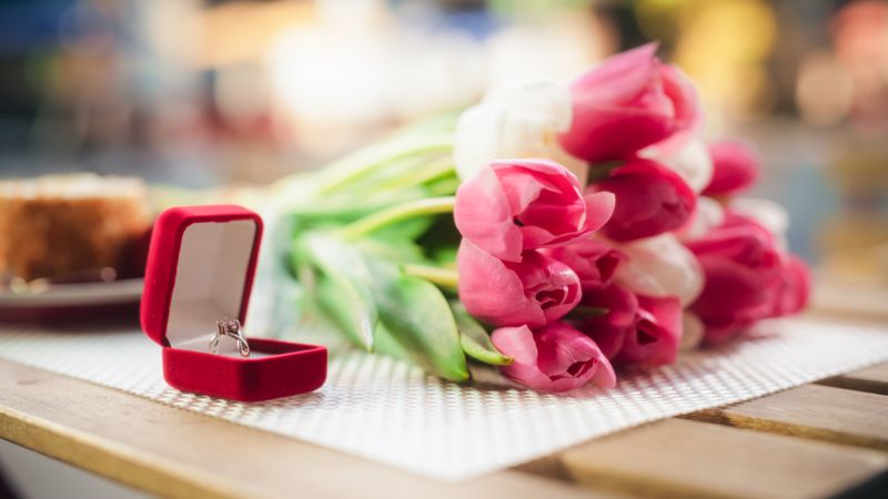 Close Up of a Beautiful Engagement Ring with a Big Gemstone Standing in a Red Box on a Table Next to Pink Tulips Bouquet. Flowers and Jewelry for a Romantic Wedding Proposal