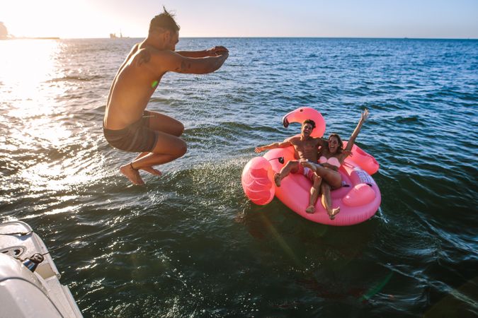 Playful male jumping midair into ocean water with friends floating on toy nearby