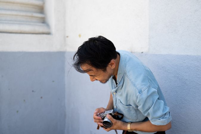 Young man laughing with head down at joke while holding a digital camera near a blue wall outdoors