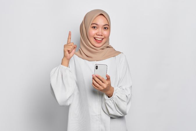Asian Muslim woman in a bright studio shoot holding cell phone and pointing upwards
