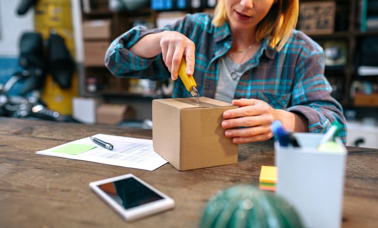 Unrecognizable female opening cardboard box with cutter. Parcel delivery and ecommerce concept.