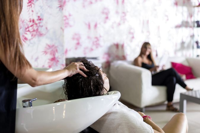 Female with hair in sink being shampooed at salon with another customer sitting in waiting area