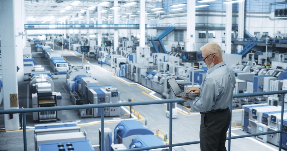 Experienced Male Engineer Standing on a Platform, Using Laptop Computer and Overlooking Production at a Modern Automated Electronics Manufacture with AI Support. Factory Executive at Work