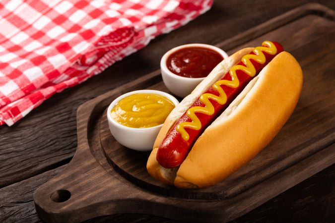 Hot dog with mustard and ketchup on wooden board and checkered napkin