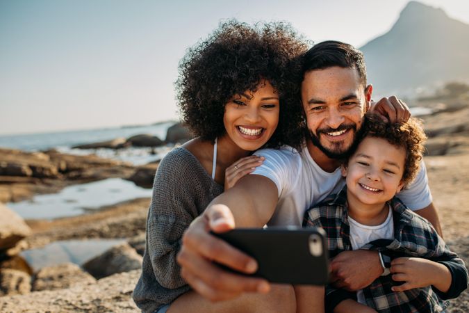 Smiling couple with kid sitting on rock near the seashore posing for a selfie