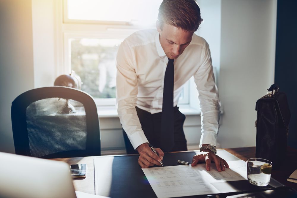 Man in shirt and tie standing over desk and writing in sunny office