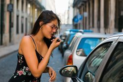 Confident woman in sunglasses applying lipstick while looking at car window on street