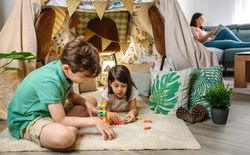 Children playing together with stacking piece game while relaxed mother reading book sitting on sofa