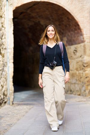 Smiling woman with camera walking away from arched passage in old town