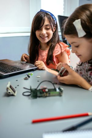 Happy female student smiling while looking to classmate assembling machine pieces