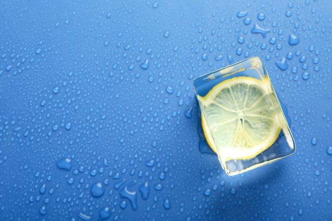 Top view of large ice cube with lemon slice on blue table, copy space