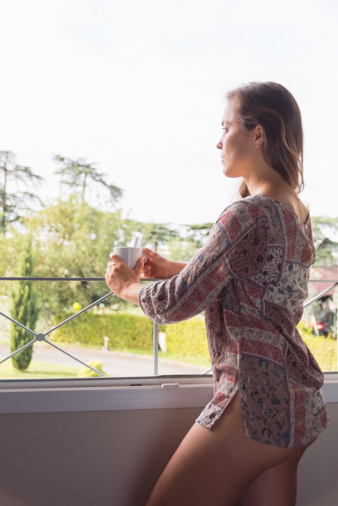 woman in a shirt with a cup of coffee or tea looking out the window in the morning