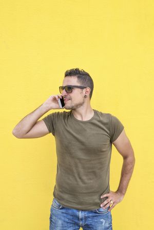 Serious male standing in front of yellow wall talking on smartphone