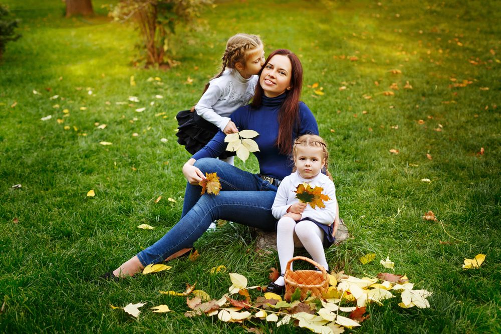 Two young blonde girls sitting with their mother in the park collecting leaves with basket