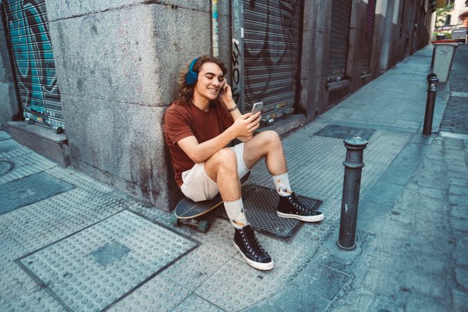 Relaxed man in headphones using smartphone while resting on longboard on street