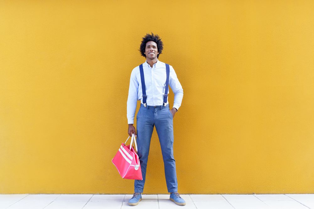 Man standing tall with curly hair wearing shirt and suspenders walking next to yellow wall