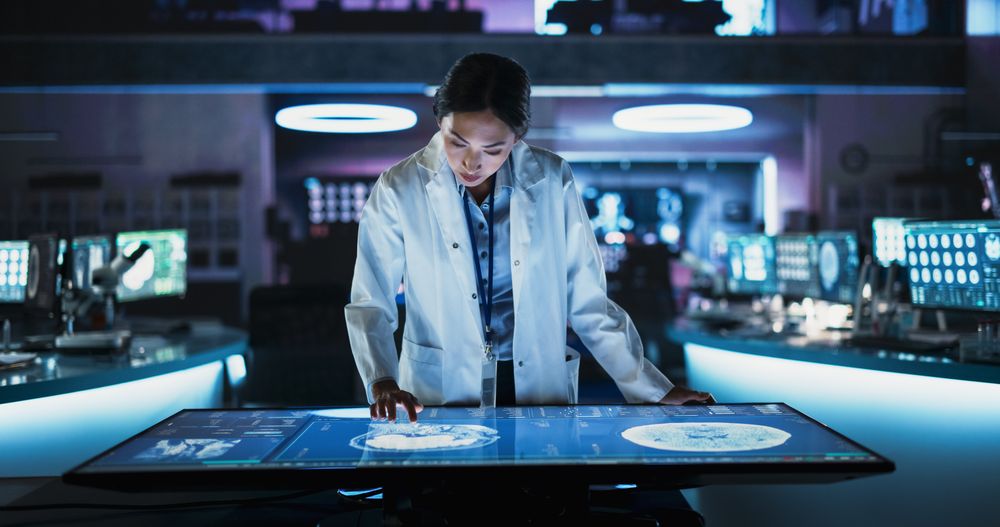 Female Asian Neuroscinetist Using Interactive Touch Screen Table With MRI Scans On Display In Modern Biotechnology Research Center.