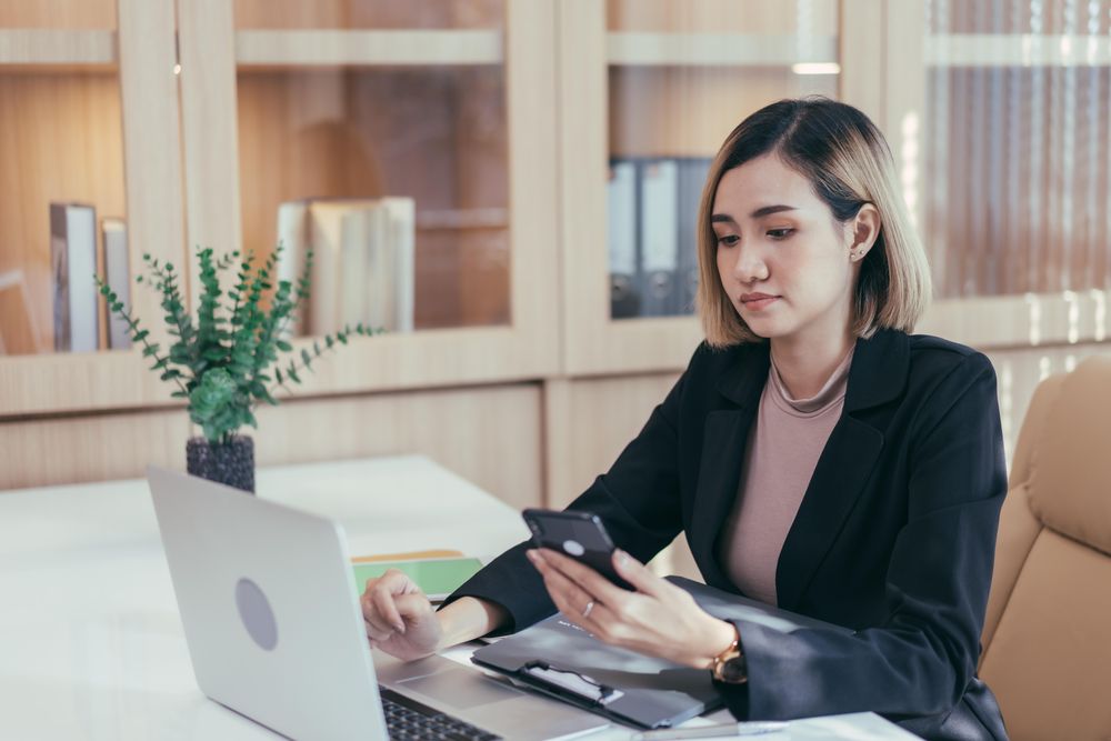 Woman checking her phone while sitting at her desk with laptop