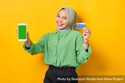 Smiling Muslim woman in headscarf and green blouse with credit card and smart phone screen mock up 56mjj0