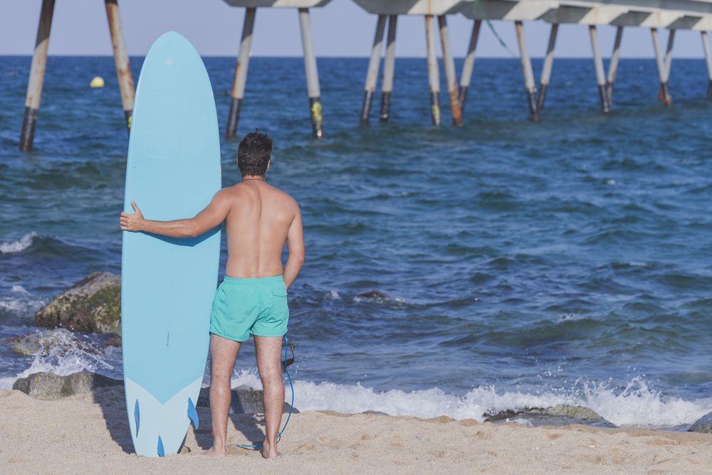 Male surfer’s back standing in the rocks on the coast near pier