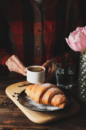 Person holding coffee, on kitchen table with large croissant on breadboard in foreground